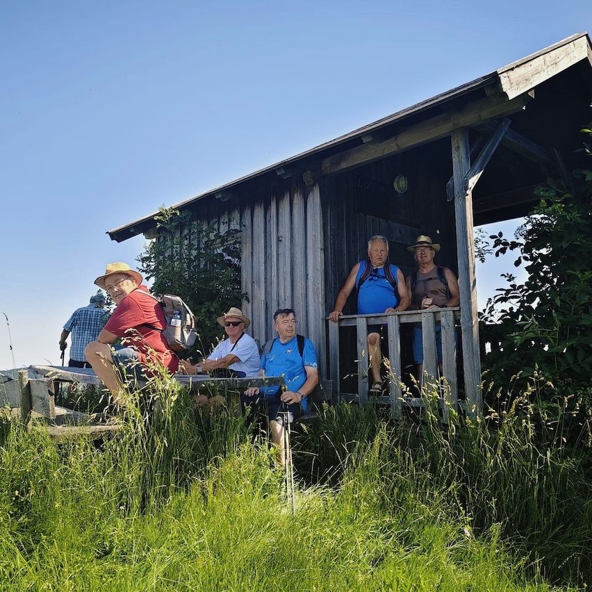 Eine Gruppe von Wanderern ruht in einer Holzhütte. Einige tragen Hüte und Rucksäcke, einer mit einem Beinprothese. Sie stehen vor einem Zaun und blicken auf hohes Gras.