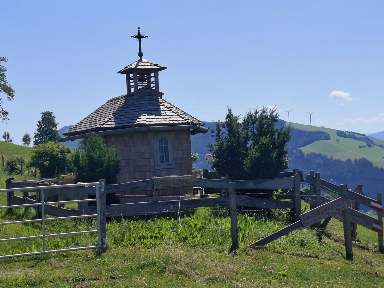 Eine kleine Kirche mit einem Kreuz auf dem Dach steht auf einem Grasfeld. In der Nähe gibt es einen Holzzaun und eine Windmühle auf den entfernten Hügeln.