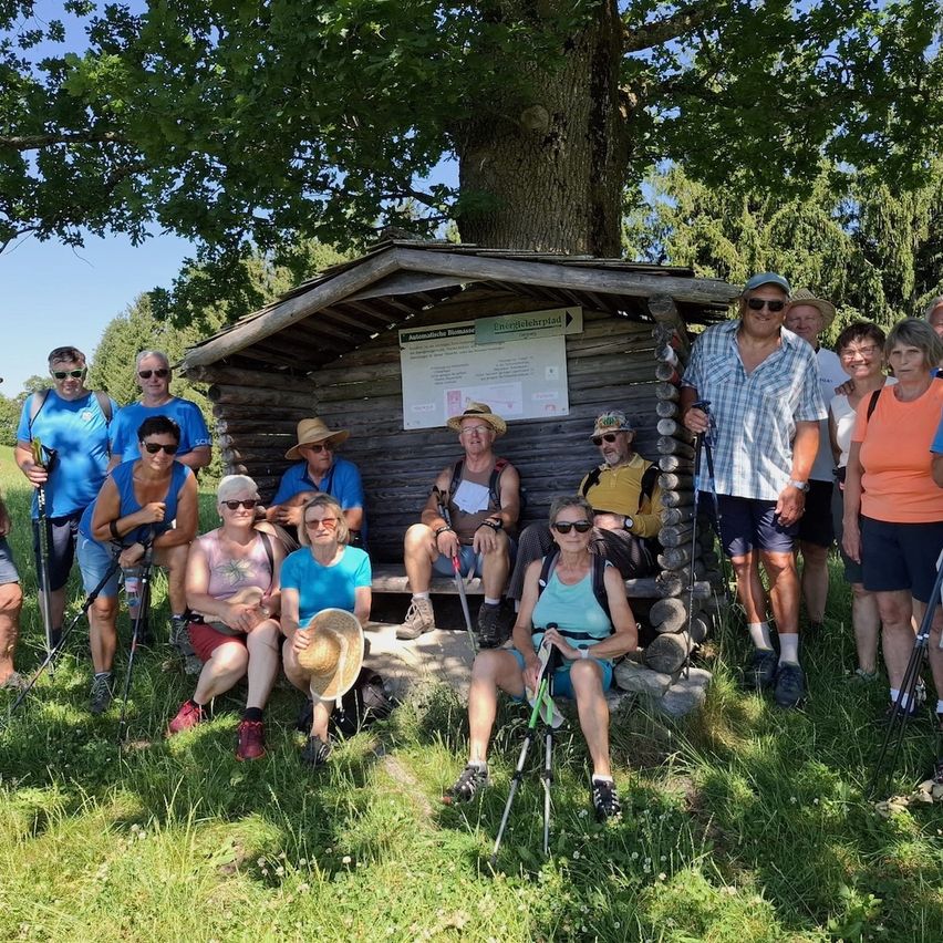 Eine Gruppe von Wanderern posiert für ein Foto neben einem Holzschuppen mit einem Schild, umgeben von Bäumen und Gras.