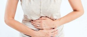Eine Frau hält sich mit angewinkelten Händen den Bauch, ihr Gesicht zeigt Anzeichen von Unbehagen. Sie trägt ein beigefarbenes T-Shirt und blaue Jeans.