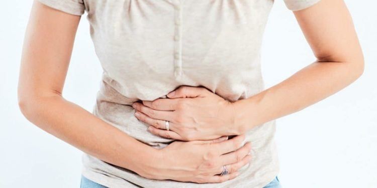 Eine Frau hält sich mit angewinkelten Händen den Bauch, ihr Gesicht zeigt Anzeichen von Unbehagen. Sie trägt ein beigefarbenes T-Shirt und blaue Jeans.