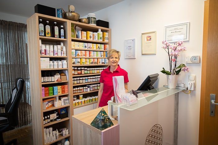 Eine Frau in einem roten Shirt steht hinter einem Empfangstresen mit einer Blumenvase. Regale mit verschiedenen Produkten stehen auf beiden Seiten, und Zertifikate hängen an der Wand.