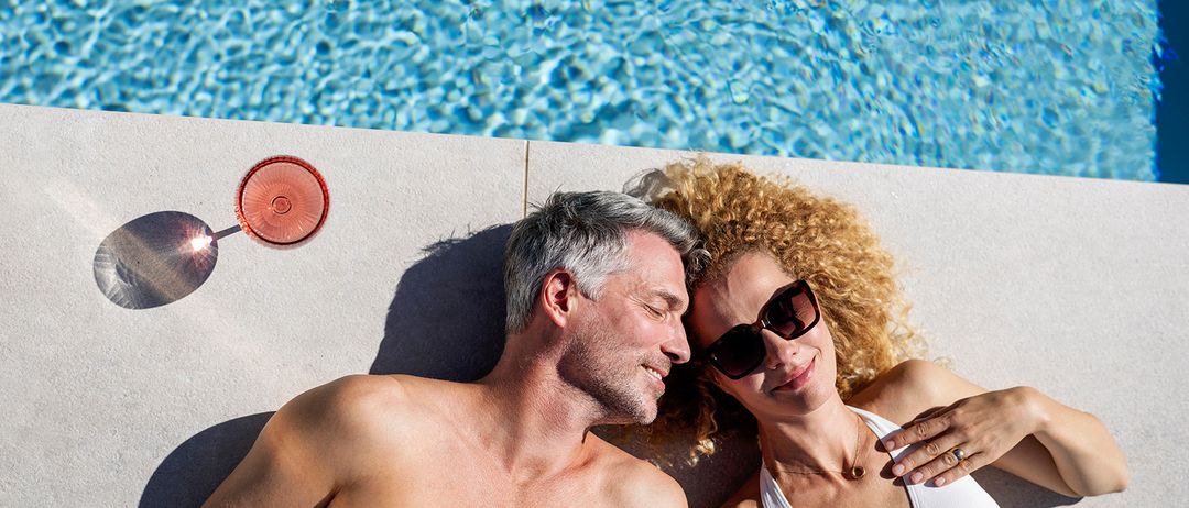 Ein Mann und eine Frau liegen am Rand eines Pools, lächeln und schauen sich an. Der Mann ist ohne Hemd, und die Frau trägt eine Sonnenbrille und eine Halskette.