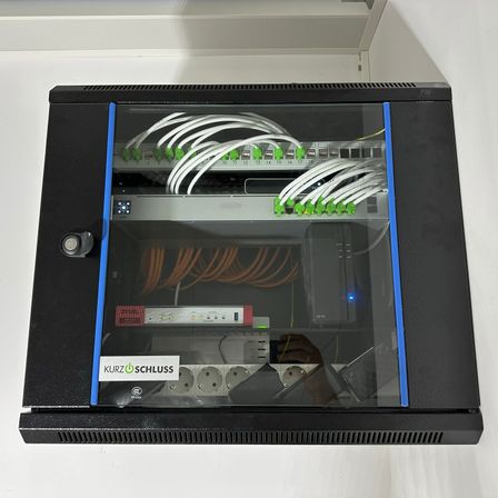 Ein schwarzer Netzwerkschrank mit Glastüren und blauen Beschlägen zeigt verschiedene Kabel und Geräte im Inneren. Auf den Geräten befinden sich Aufkleber wie 'Kurz Schluss' und 'ZyXEL'.