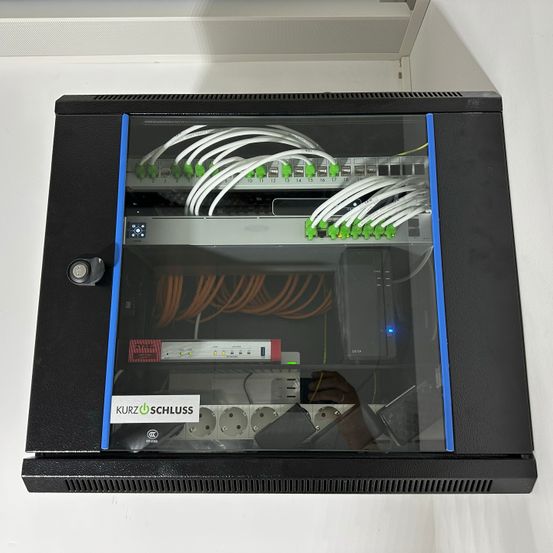 Ein schwarzer Netzwerkschrank mit Glastüren und blauen Beschlägen zeigt verschiedene Kabel und Geräte im Inneren. Auf den Geräten befinden sich Aufkleber wie 'Kurz Schluss' und 'ZyXEL'.