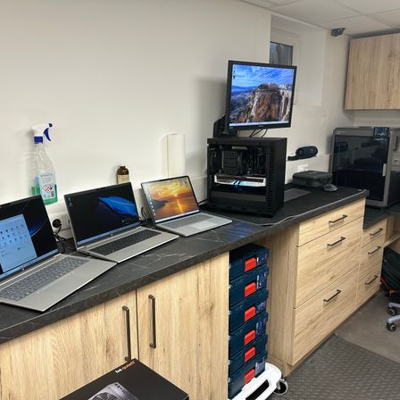 Ein technisches Setup mit mehreren Laptops, einem Monitor und einem Computergehäuse auf einer Arbeitsplatte. Verschiedene Werkzeuge und eine Sprühflasche sind ebenfalls vorhanden.