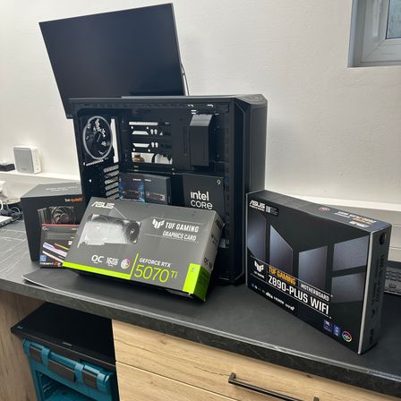 Ein Computeraufbau auf einem Schreibtisch umfasst ein offenes PC-Gehäuse, einen Monitor und mehrere Kartons, darunter einer für einen Intel Core-Prozessor und ein weiterer für eine TUF Gaming-Grafikkarte.