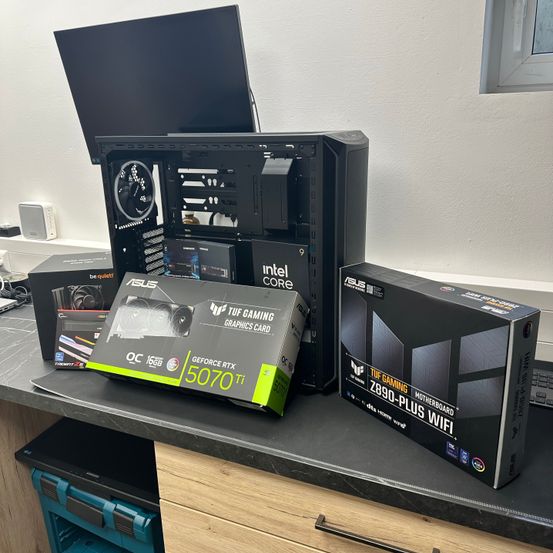 Ein Computeraufbau auf einem Schreibtisch umfasst ein offenes PC-Gehäuse, einen Monitor und mehrere Kartons, darunter einer für einen Intel Core-Prozessor und ein weiterer für eine TUF Gaming-Grafikkarte.
