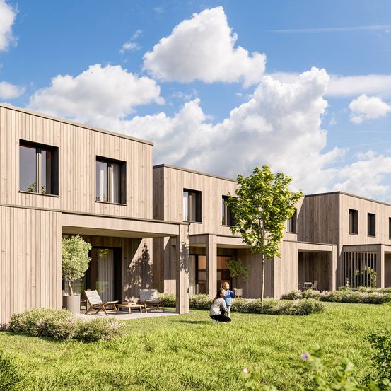 Drei modulare Häuser mit Holzfassaden in einer Wohngegend. Eine Familie sitzt im Garten, und ein junges Mädchen spielt mit einem Hund. Draußen stehen Stühle und ein Tisch, und das Haus hat große Fenster. Der Himmel ist blau mit weißen Wolken.