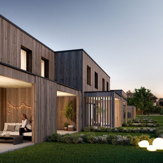 Modernes Holzhaus mit Terrasse und einer Frau, die auf einer Couch unter Lichterketten sitzt.