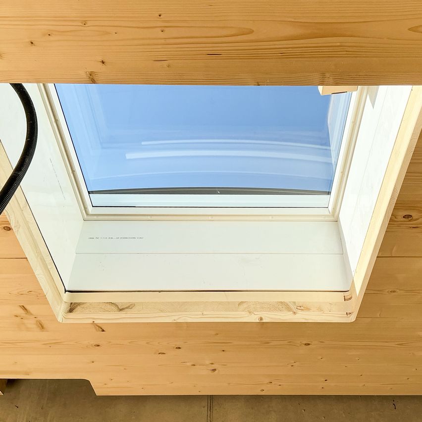 A wooden attic window with a blue sky visible through the glass. The frame is also wooden, and it is installed on a wooden ceiling.