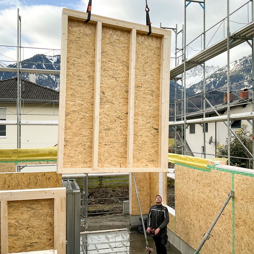 A man in a construction site is looking up at a large wooden panel being lifted by a crane. Behind him is a building under construction with scaffolding.