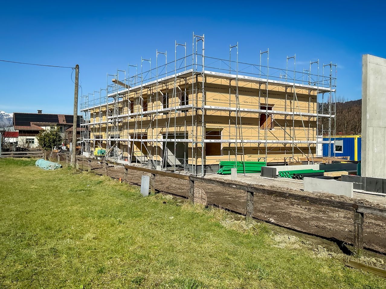 A building under construction with scaffolding. It has multiple floors, windows, and an open doorway. Nearby, there are green poles and a fence.