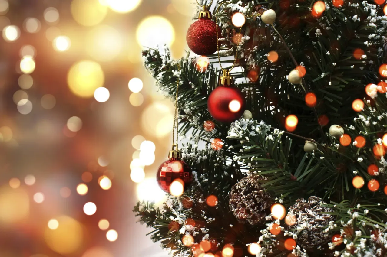 Ein Weihnachtsbaum mit roten Kugeln, Schnee und leuchtenden Lichtern in einem festlichen Ambiente. Der Baum ist von unscharfen gelben Lichtern umgeben.