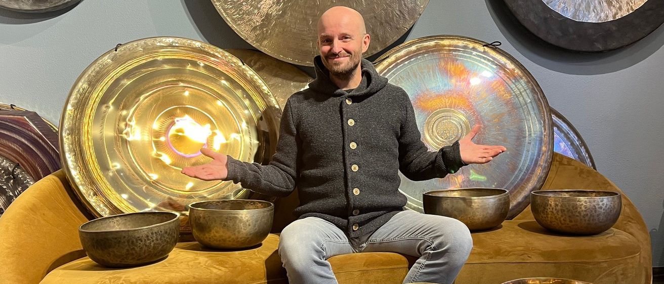 Ein kahlköpfiger Mann sitzt mit ausgestreckten Armen unter goldenen Gongs und Schalen auf einem Samtsofa, umgeben von beleuchteten Gongs an der Wand.