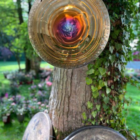 Ein kreisförmiges Glasobjekt mit einem leuchtenden, mehrfarbigen Zentrum hängt von einem Baum in einem Garten. Grüne Blätter bedecken den Baum, und im Hintergrund sind rosa Blumen zu sehen.