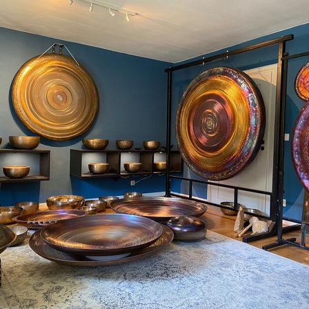 Ein Raum mit blauen Wänden zeigt verschiedene Musikinstrumente, darunter hängende Gongs und Schüsseln auf einem Holzboden. Im Vordergrund befindet sich ein Teppichboden.