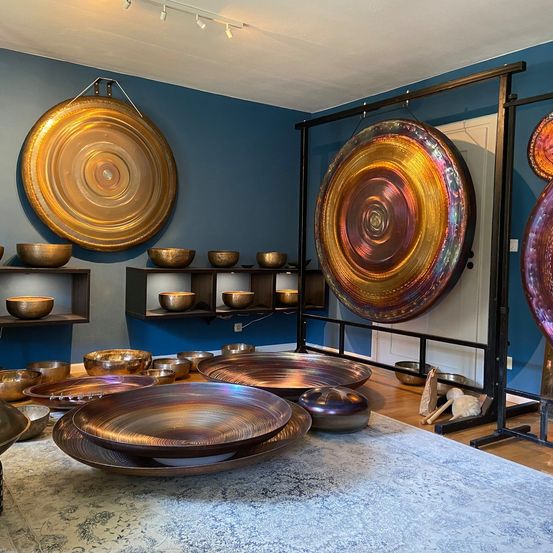 Ein Raum mit blauen Wänden zeigt verschiedene Musikinstrumente, darunter hängende Gongs und Schüsseln auf einem Holzboden. Im Vordergrund befindet sich ein Teppichboden.