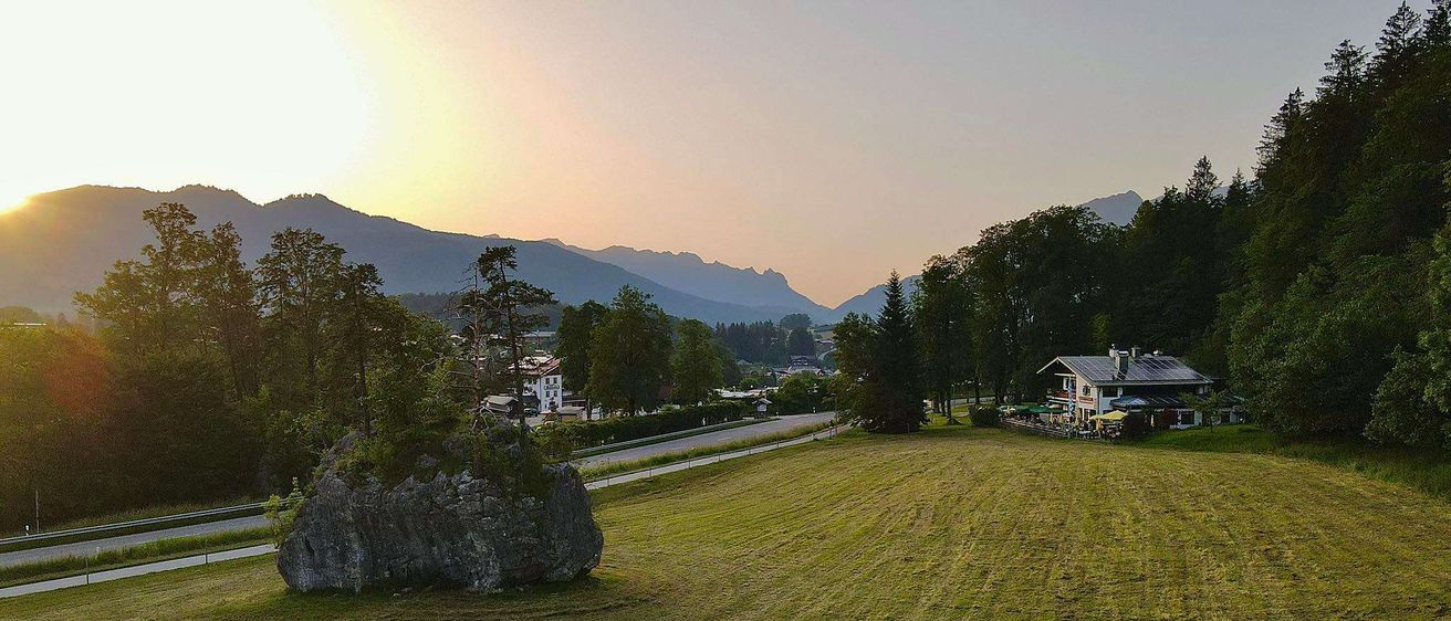 Eine landschaftliche Ansicht einer ländlichen Gegend mit Bergen im Hintergrund, einem großen Felsbrocken im Vordergrund und einem Gebäude mit Solarpanelen auf der rechten Seite.