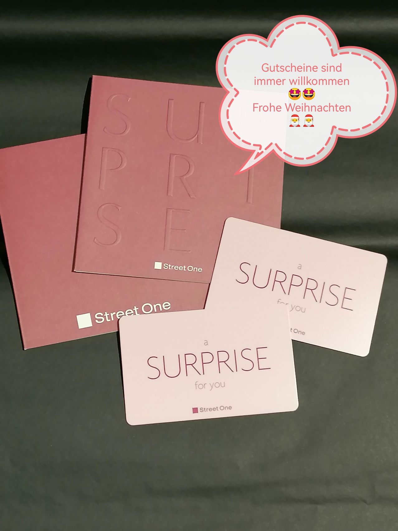 Drei rosafarbene Street One Geschenkkarten sind auf einem schwarzen Hintergrund ausgestellt. Die oberste Karte hat eine Sprechblase mit Emojis. Die mittlere Karte sagt 'eine Überraschung für Sie'. Die unterste Karte liest 'ÜBERRASCHUNG für Sie'.