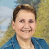 Eine Frau mit kurzen Haaren, die eine Jeansjacke und ein weißes Hemd trägt, lächelt vor einem unscharfen Hintergrund von Bäumen.