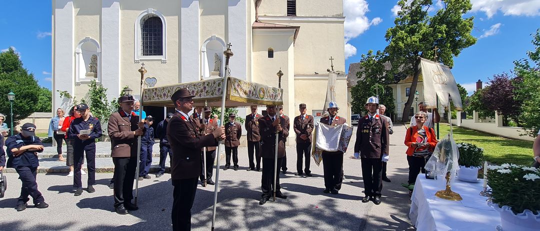 Eine Gruppe uniformierter Personen steht in einer Reihe vor einer Kirche und hält ein dekoriertes Baldachin. Hinter ihnen beobachten andere, und zwei Personen sitzen an einem Tisch mit einer Blumenarrangement.