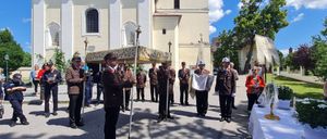 Eine Gruppe uniformierter Personen steht in einer Reihe vor einer Kirche und hält ein dekoriertes Baldachin. Hinter ihnen beobachten andere, und zwei Personen sitzen an einem Tisch mit einer Blumenarrangement.