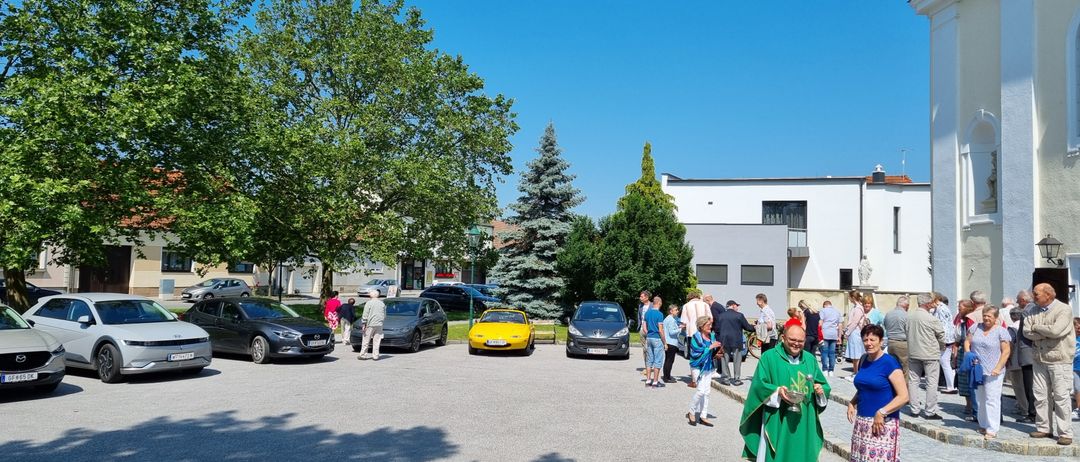 Ein Priester in einem grünen Gewand geht auf einem Parkplatz. Autos sind in der Nähe geparkt, und Menschen stehen in Gruppen. Bäume und Gebäude sind im Hintergrund.
