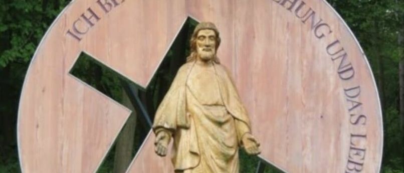 Ein hölzernes Schild mit einer Statue von Jesus in der Mitte, umgeben von einem Kreuz und einem deutschen Satz.