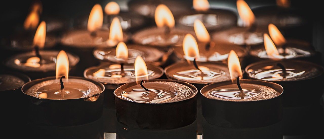 Mehrere kleine Kerzen mit schwarzen Sockeln und weißer Wachs brennen in einem dunklen Bereich. Die Flammen sind entzündet und etwas Wachs ist geschmolzen.