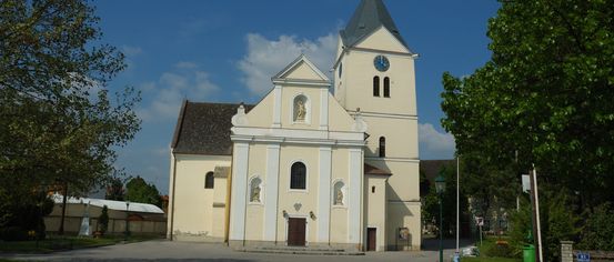 Eine Kirche mit Glockenturm befindet sich in einer ruhigen Gegend mit einem Grasfeld, Bäumen und einer Straßenlaterne. Sie verfügt über ein Kreuz auf der Spitze, Statuen und Bogenfenster.