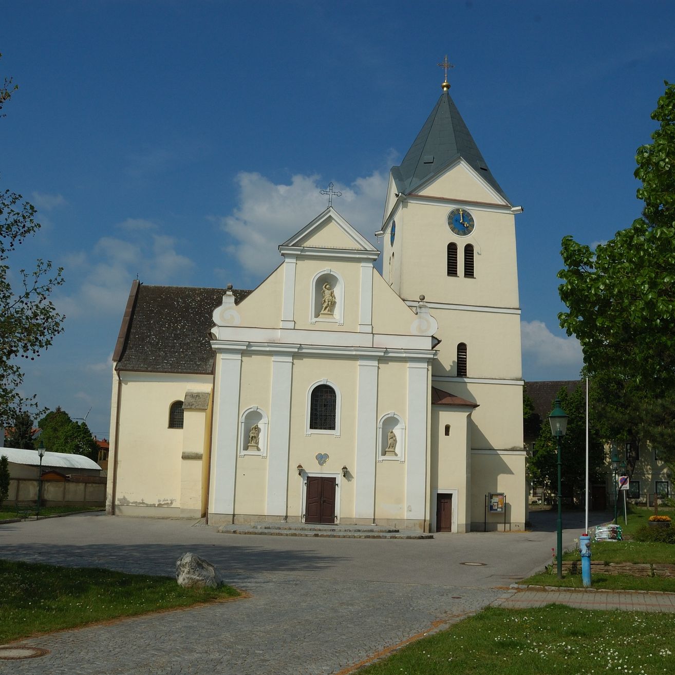 Eine Kirche mit einem hohen Turm, der eine Uhr zeigt, steht vor einem Parkplatz. Bäume und Straßenlaternen flankieren das Gebäude.