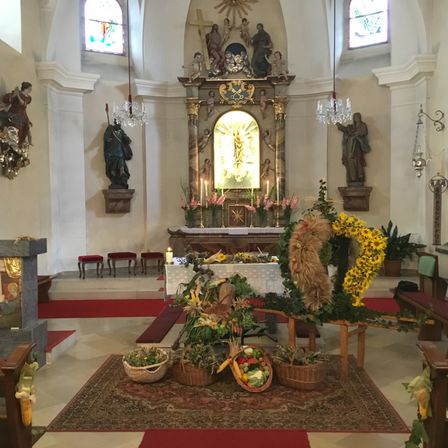 Ein Kirchenaltar mit Kerzen und Blumen. An den Wänden befinden sich Statuen. Der Boden ist mit einem roten Teppich und Blumen bedeckt.