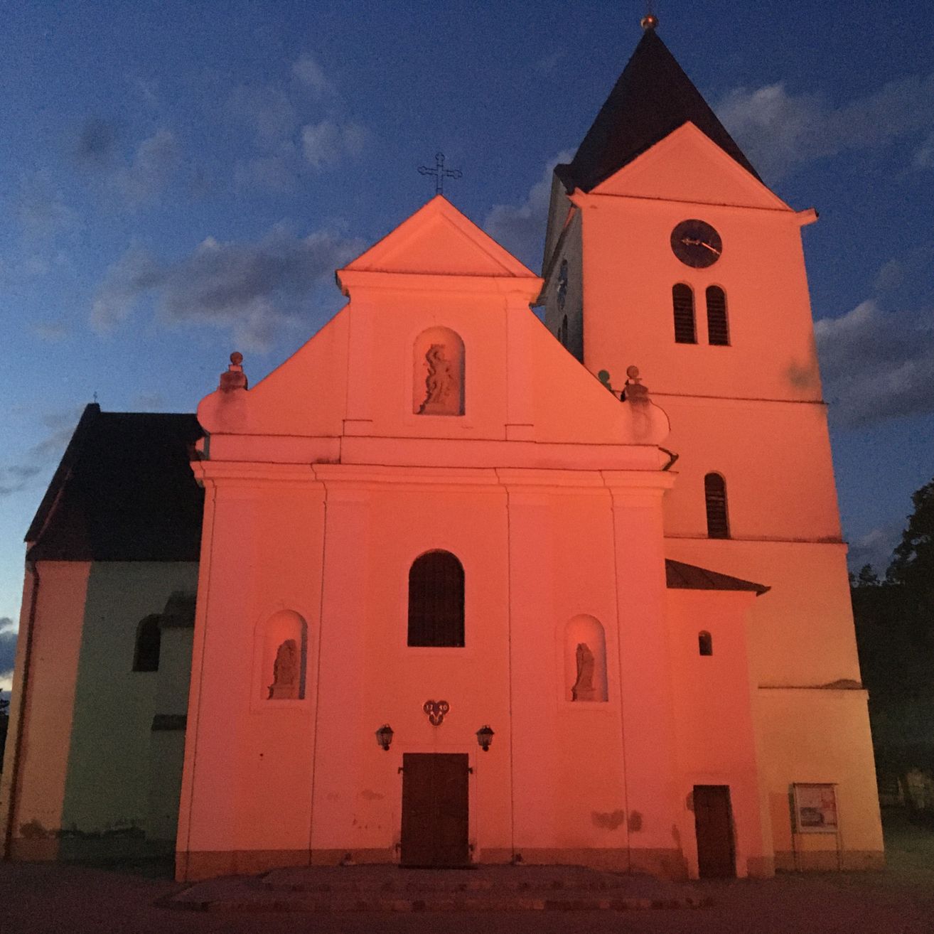 Eine Kirche mit roter Fassade steht in der Dämmerung. Das Gebäude hat einen hohen Turm mit einer Uhr und Kreuzen oben. Es gibt Statuen in gewölbten Nischen.
