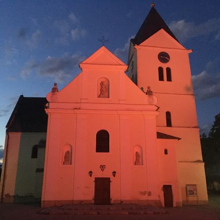 Eine Kirche mit roter Fassade steht in der Dämmerung. Das Gebäude hat einen hohen Turm mit einer Uhr und Kreuzen oben. Es gibt Statuen in gewölbten Nischen.