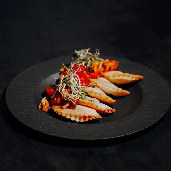 Ein schwarzer Teller zeigt sechs Ravioli, gefüllt mit roter Sauce und belegt mit Kirschtomaten, Microgreens und Pilzen.