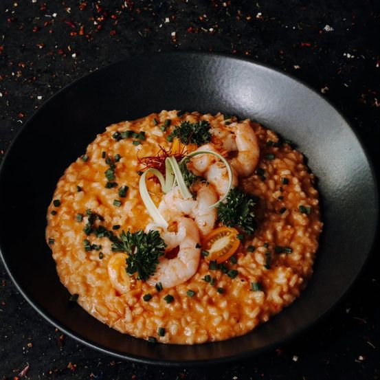 Ein schwarzer Teller hält ein orangefarbenes Risotto-Gericht mit Garnelen, Frühlingszwiebeln und Petersilie auf einem dunklen Tisch.