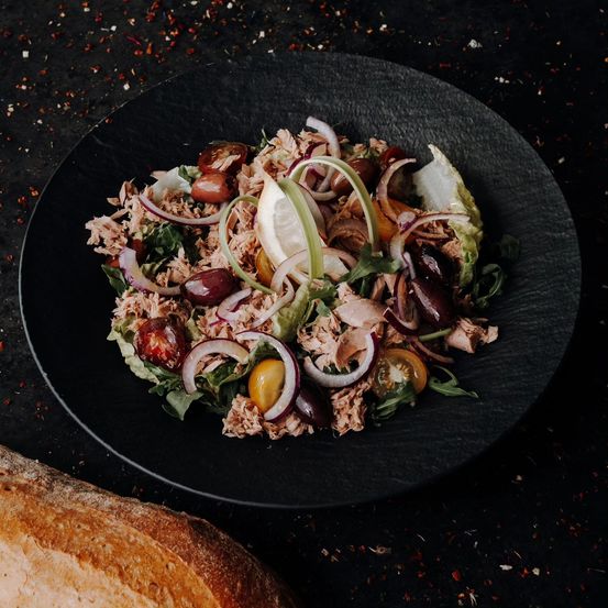 Ein schwarzer Teller mit einem Thunfischsalat, garniert mit Zwiebeln, Tomaten, Oliven und Zitronenscheiben, begleitet von einem Stück Brot.