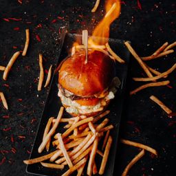 Ein brennender Burger mit Pommes auf einem schwarzen Teller, umgeben von verstreuten Pommes und roten Chiliflocken.
