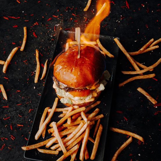 Ein brennender Burger mit Pommes auf einem schwarzen Teller, umgeben von verstreuten Pommes und roten Chiliflocken.