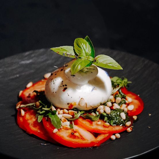 Ein Teller mit Burrata-Käse, roten Tomaten, Basilikumblättern und Pinienkernen auf einer schwarzen Oberfläche.