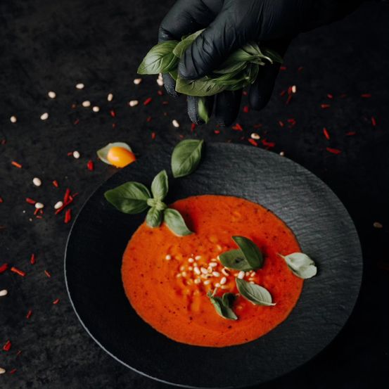 Eine Nahaufnahme der Hand eines Kochs, die frische Basilikumblätter über eine Schüssel Tomatensuppe hält, garniert mit gehackten Pinienkernen und Basilikum.