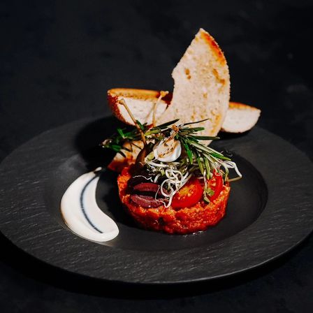 Auf einem schwarzen Teller befindet sich ein rotes Gericht mit Tomaten und Kräutern, garniert mit einer weißen Sauce und zwei Brotscheiben.