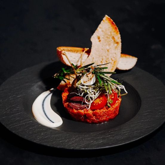 Auf einem schwarzen Teller befindet sich ein rotes Gericht mit Tomaten und Kräutern, garniert mit einer weißen Sauce und zwei Brotscheiben.