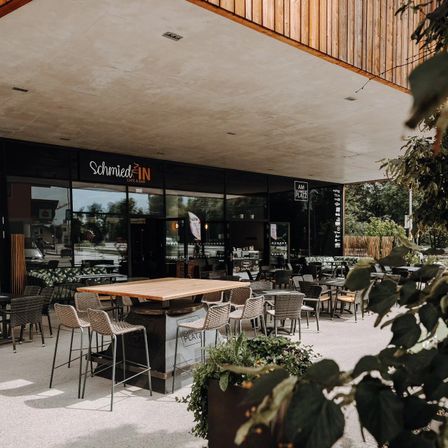Außensitzplätze im Schmied IN, einem modernen Cafe mit Holzakzenten, bieten Tische und Stühle unter einem Vordach. Die Glasfassade spiegelt die Umgebung wider, und ein Schild lautet 'Schmied IN'.