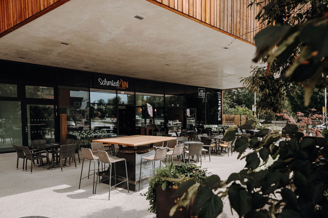 Ein moderner Außensitzbereich eines Restaurants namens Schmied IN. Tische und Stühle sind in einem offenen Bereich mit einem Holztisch und einer Glaswand angeordnet.