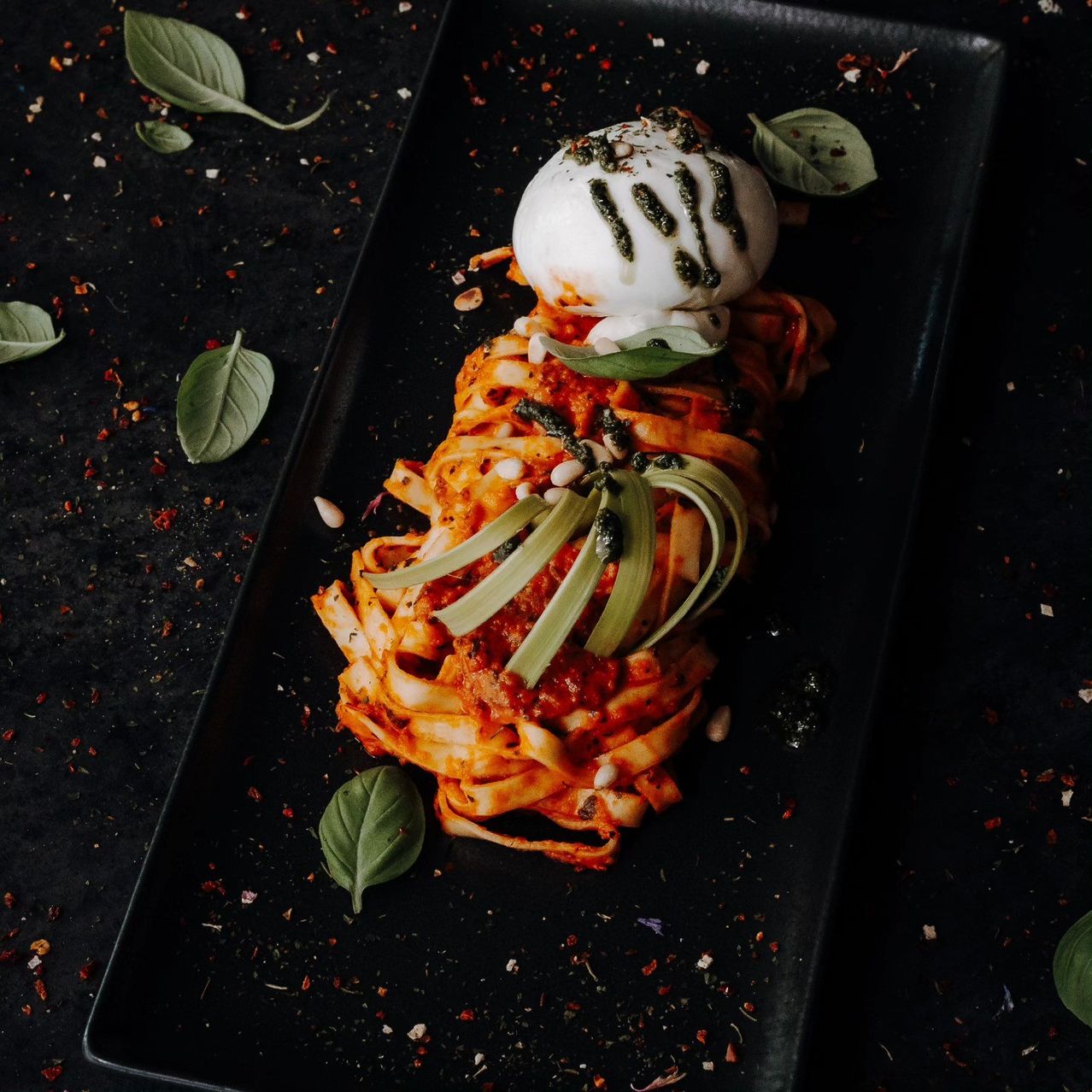 Ein Teller mit Pasta, überzogen mit Tomatensauce, Basilikum und einer Mozzarella-Kugel auf einer schwarzen Oberfläche.