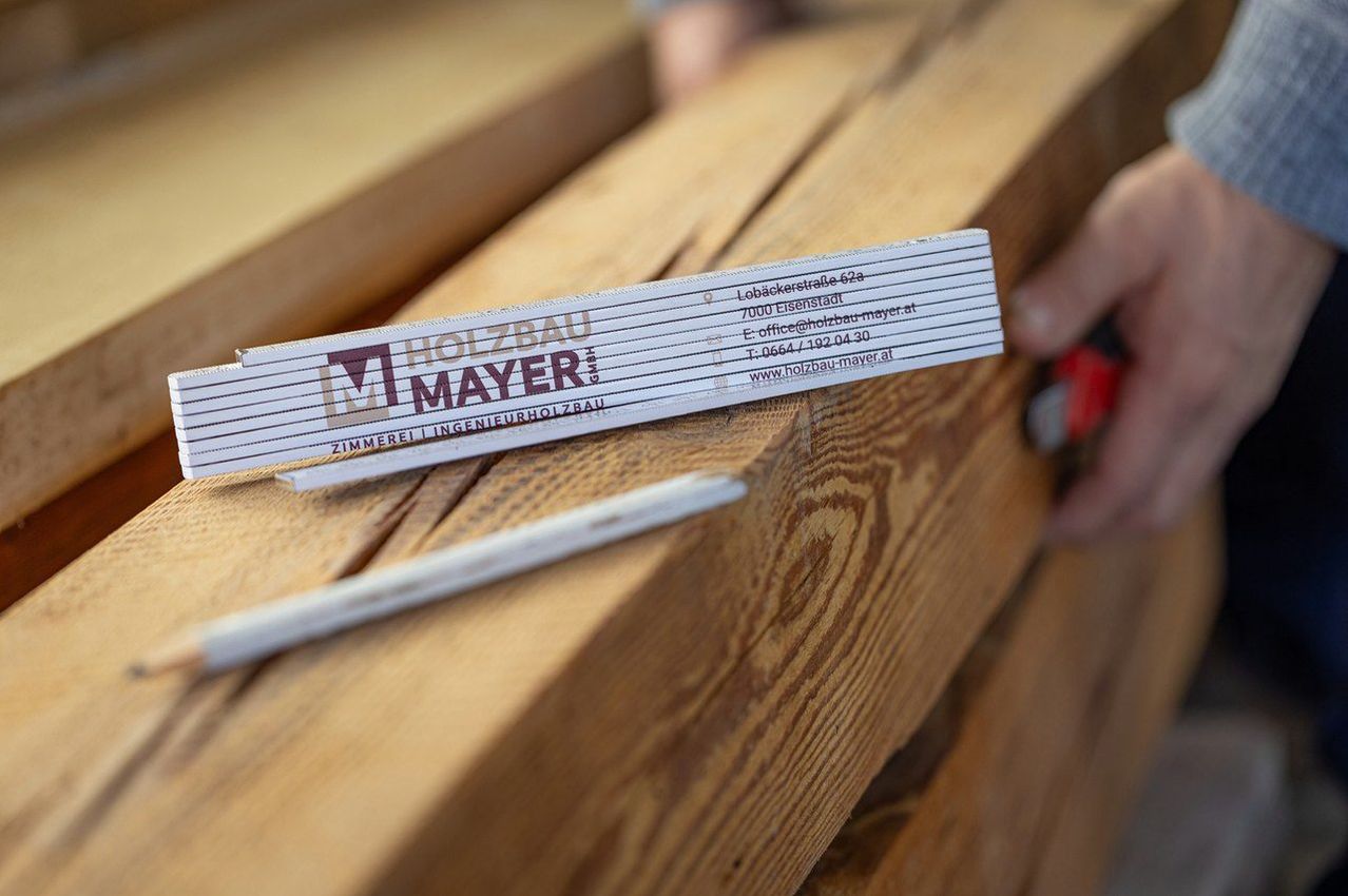 A close-up of a person's hand holding a piece of paper with the text 'Holzbau Mayer'. The paper is placed on a wooden surface. A small wooden stick is nearby.