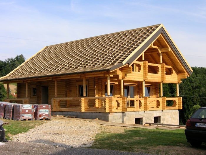 A wooden house with a tiled roof and wooden porch is under construction with materials on the ground. A car is parked nearby.