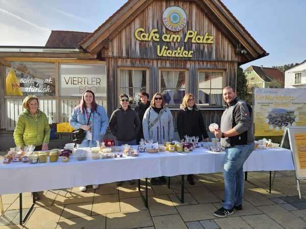 Sieben Personen, darunter zwei Frauen, stehen vor einem Tisch voller Lebensmittel. Dahinter ist ein Gebäude mit den Worten 'Cafe am Platz Victler' zu sehen.